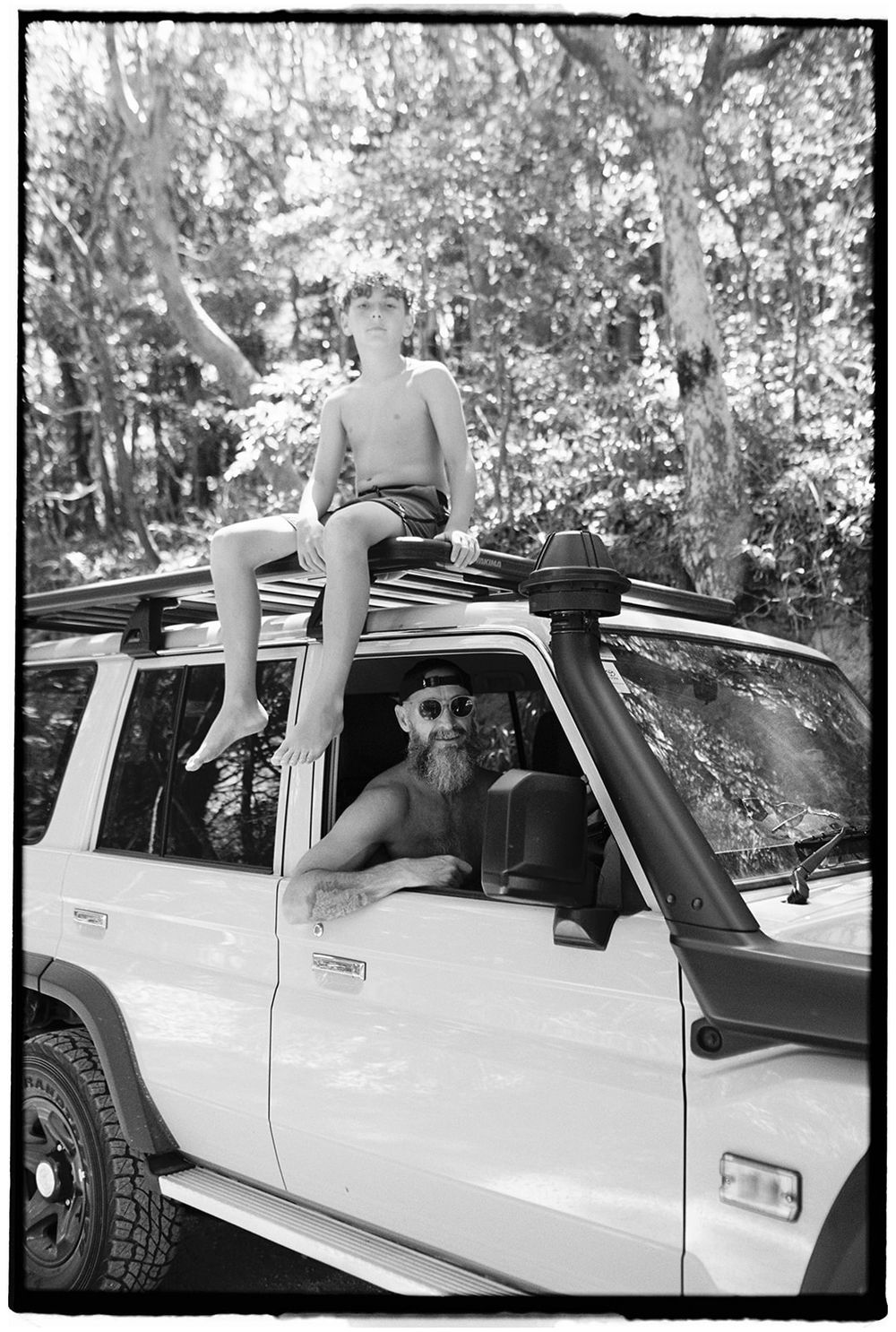 Black and white photo of bearded man in SUV with child sitting on roof rack, surrounded by trees.