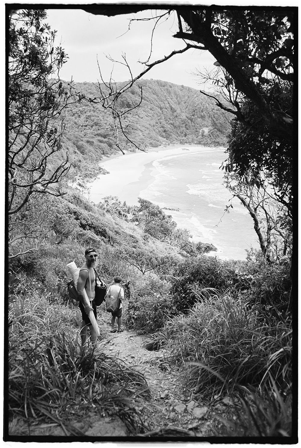 Black and white photo of two hikers on a coastal trail with lush hills and a scenic beach visible below.