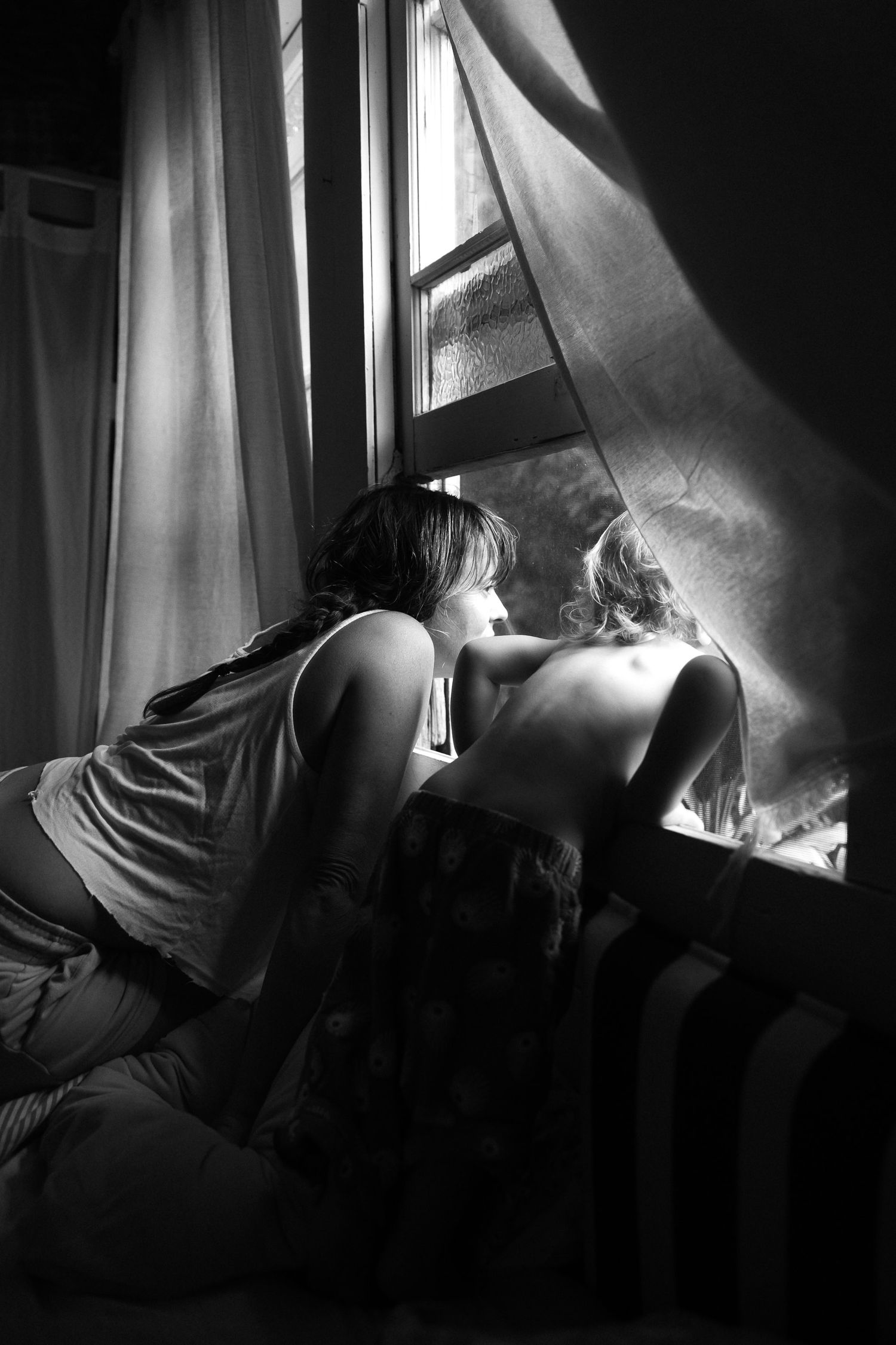 Two figures lean towards a window with billowing curtains in a moody black and white photograph.