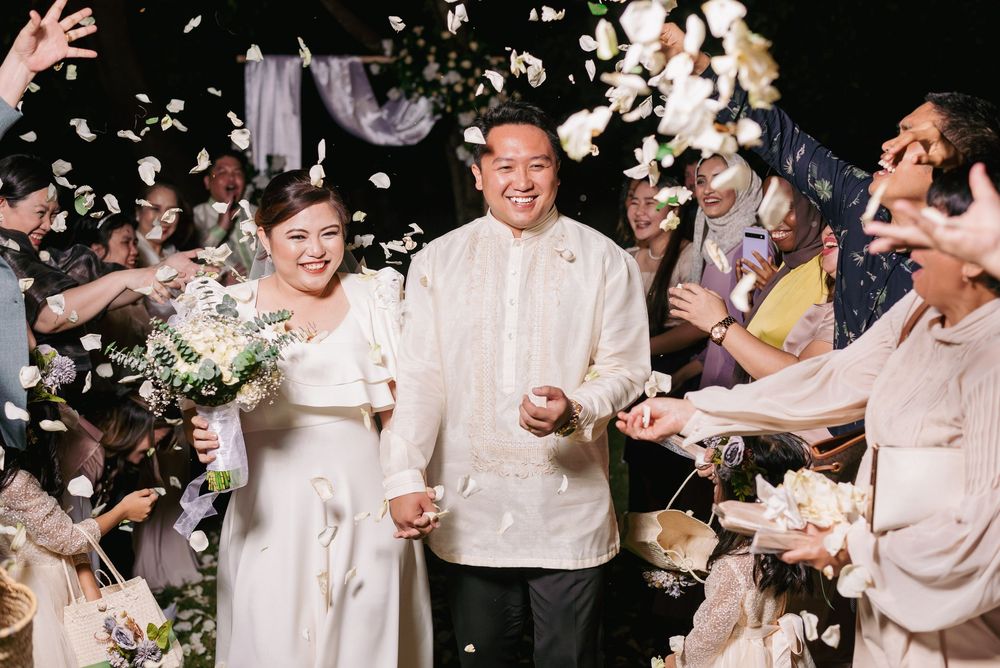 Newlyweds walk through shower of flower petals during wedding recessional while wearing traditional Filipino barong attire.