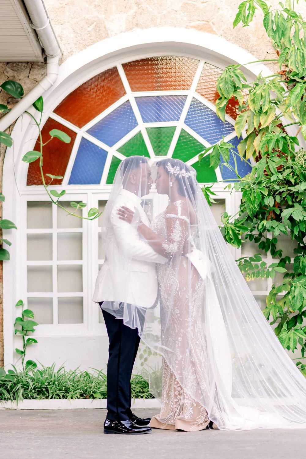 Wedding couple embracing under colorful stained glass window with flowing veil.
