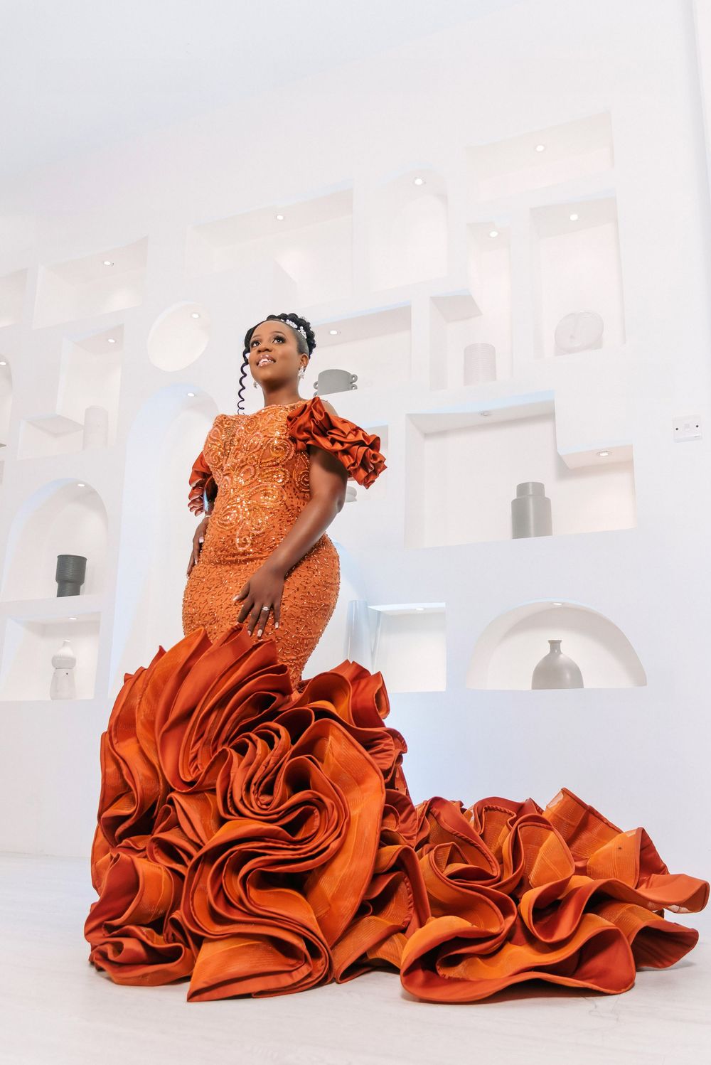 Person in flowing orange formal gown with dramatic ruffles posing in white room.
