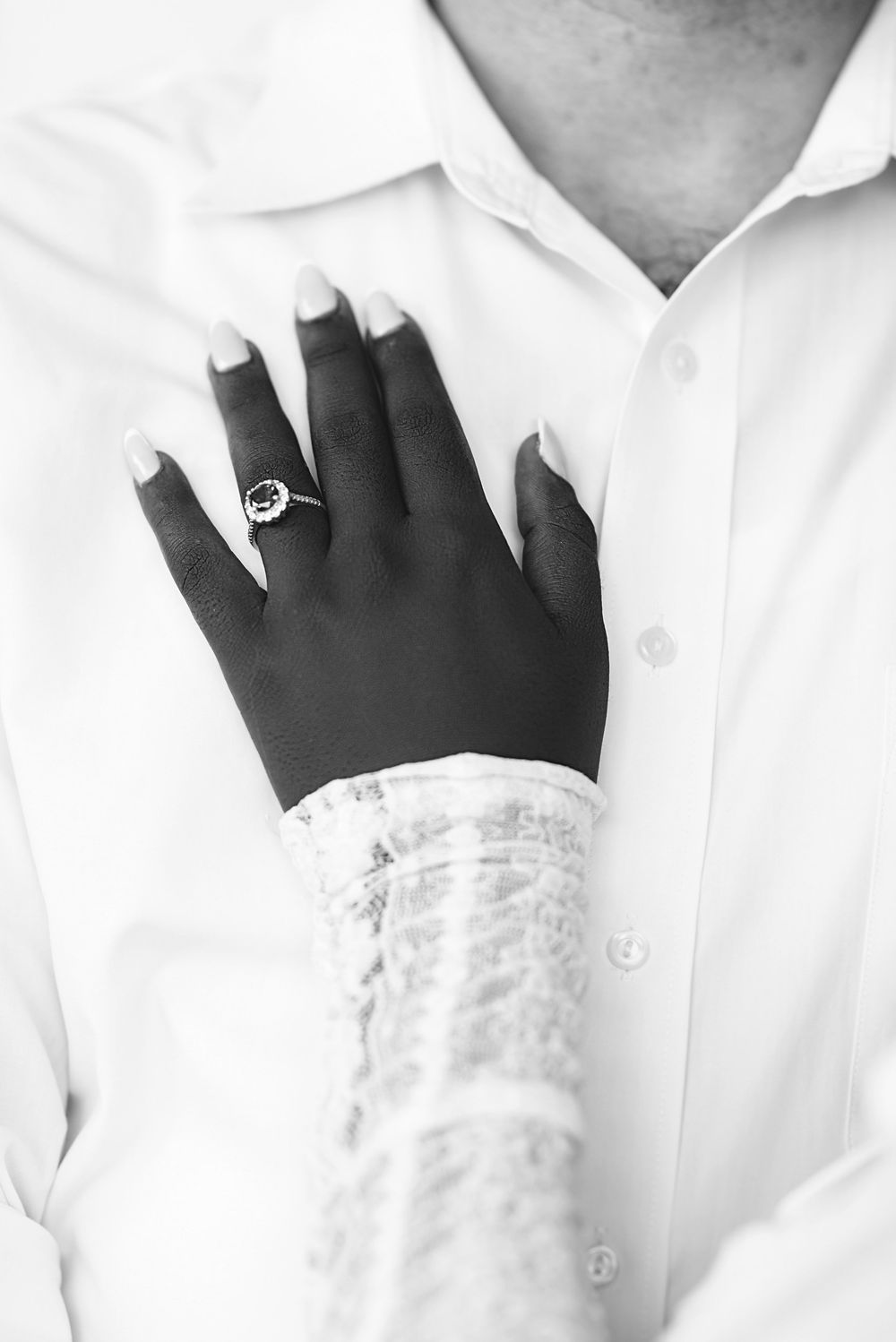Black and white close-up of hands wearing engagement ring with delicate lace sleeve and white shirt details.