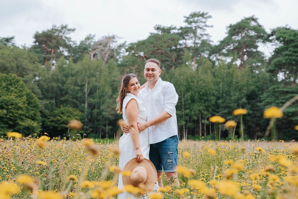 A couple holds hands while walking through a vibrant yellow wildflower field surrounded by tall pine trees.