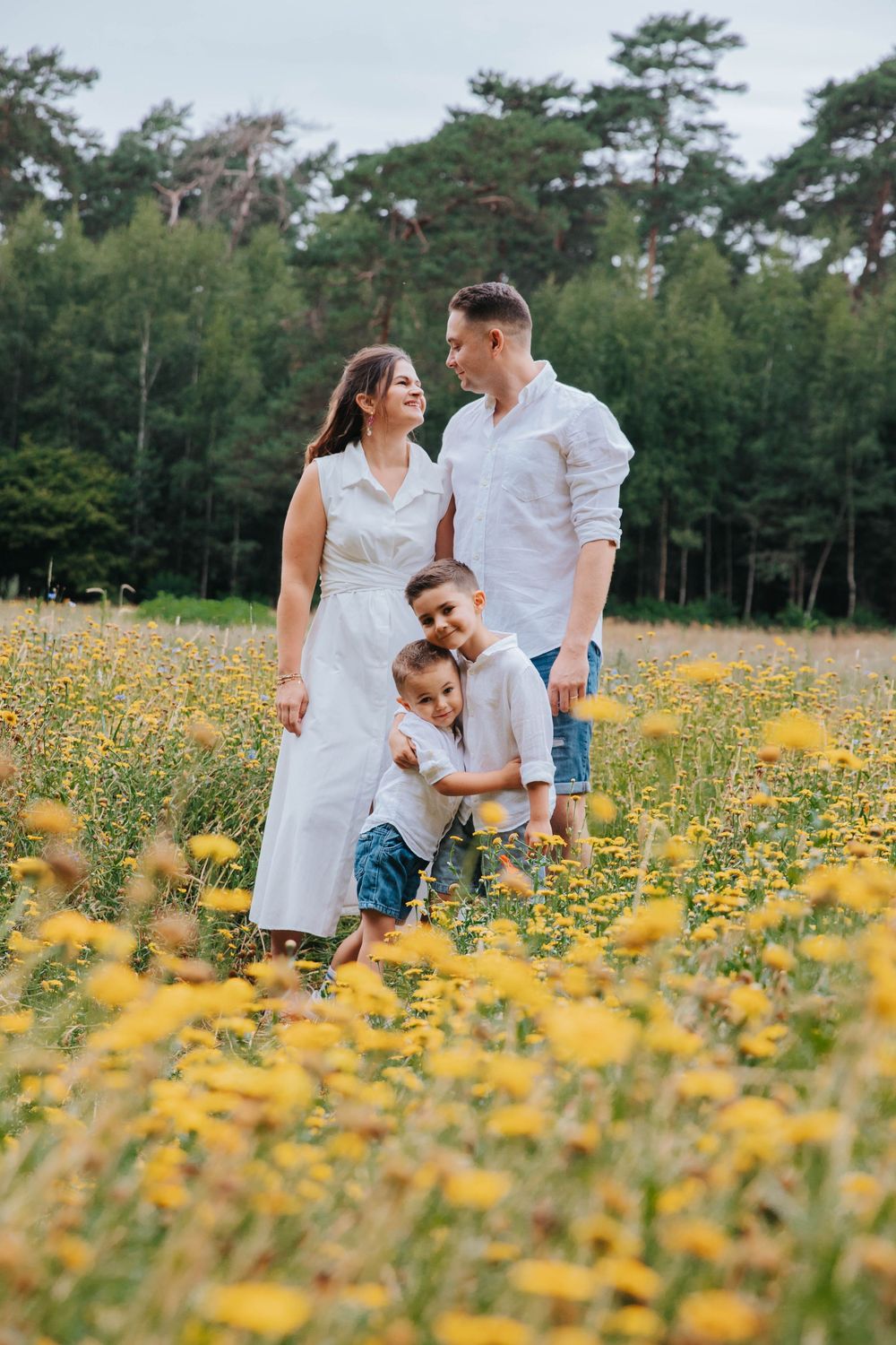 A happy family stands together in a sunny meadow filled with bright yellow wildflowers against a backdrop of evergreen trees.
