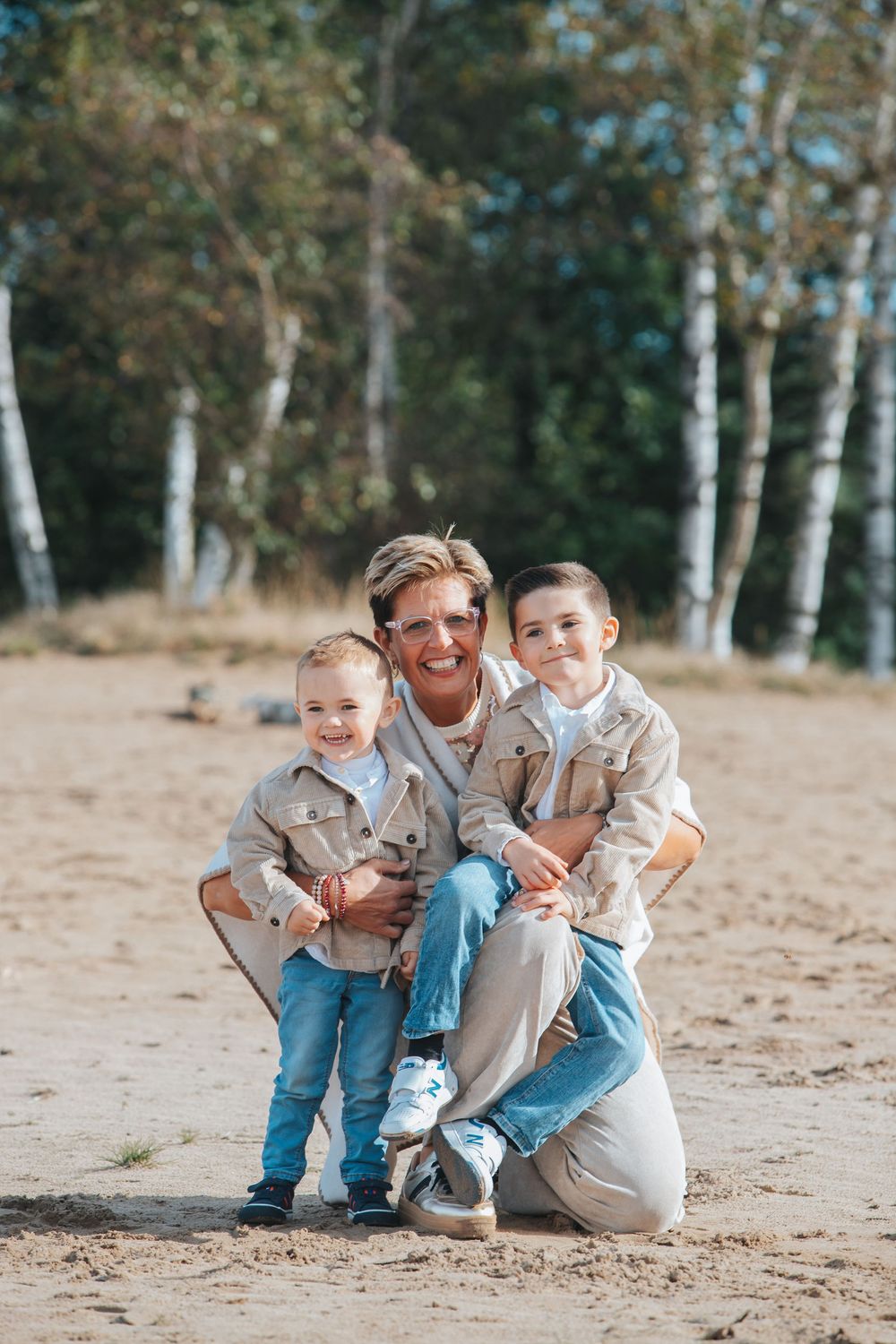 Three people wearing blue jeans and light colored tops pose together on a sandy beach with birch trees in background.