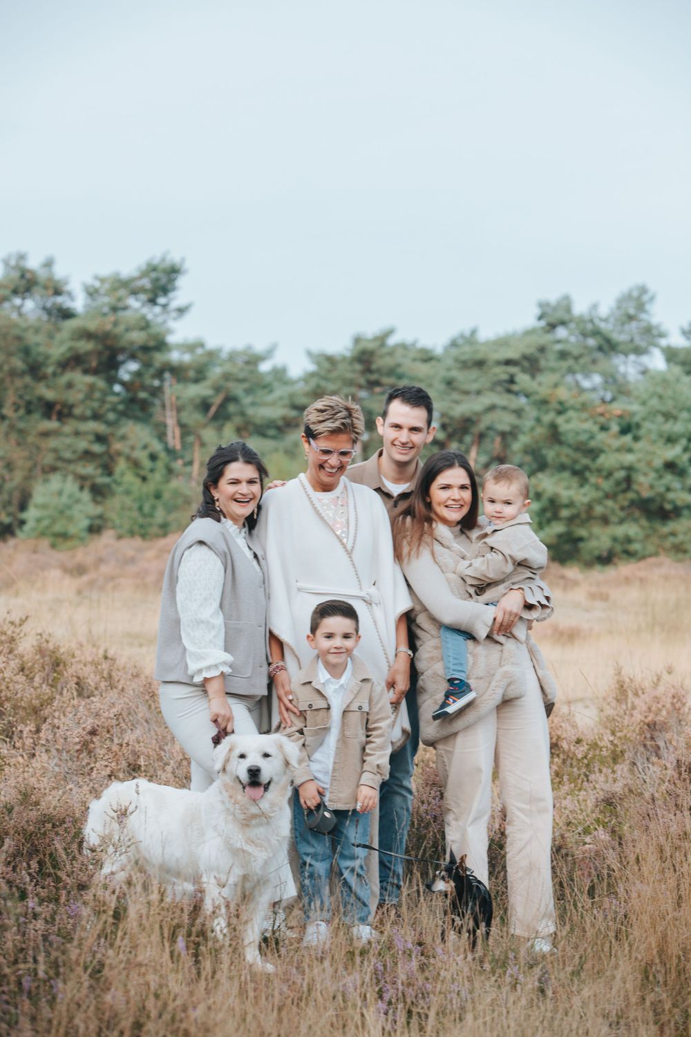 A happy family in neutral-colored clothing poses together with their white dog in a rustic field setting.