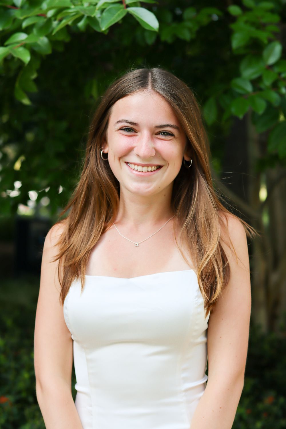Smiling portrait series in white sleeveless top with natural outdoor greenery backdrop.