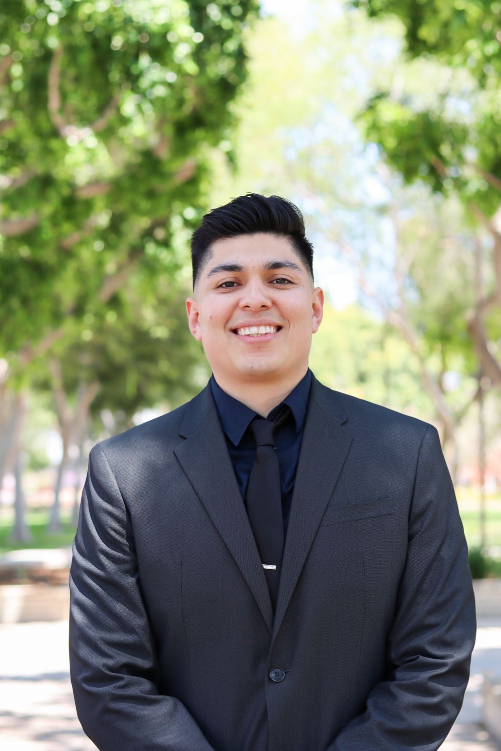 Professional portrait series in formal black suit and tie against natural outdoor background with trees.