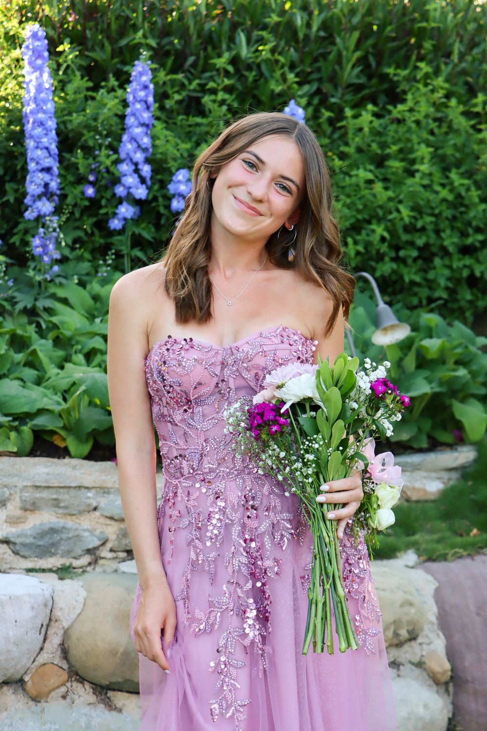 Person in pink formal dress holding colorful flower bouquet against garden backdrop with purple flowers.