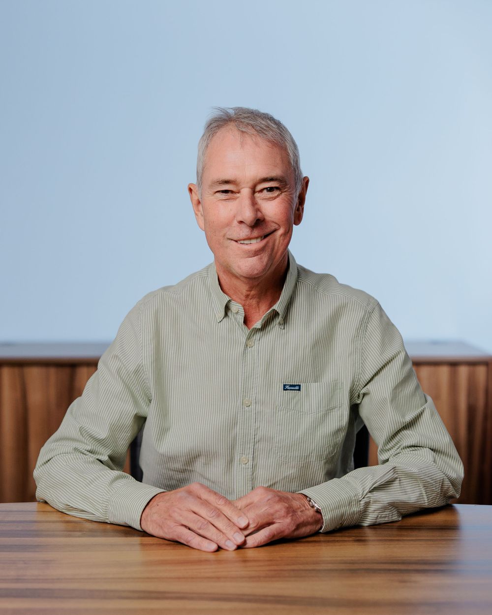 Professional in light green button-down shirt seated at wooden desk with wood panel background.