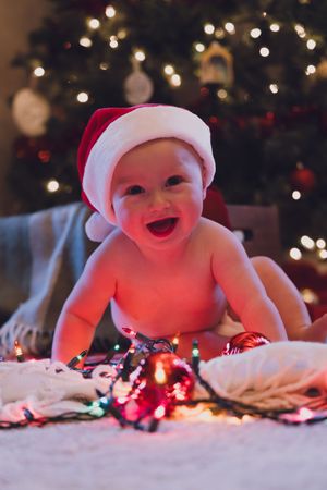 Baby lying on white blanket with glowing Christmas lights and bokeh background creates festive holiday scene.
