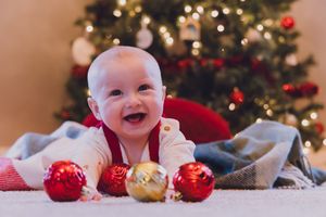 A baby smiles joyfully while sitting in front of a decorated Christmas tree with red and gold ornaments.