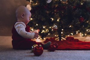 A baby in festive holiday attire sits by a decorated Christmas tree with twinkling lights and ornaments.