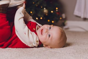Baby in red holiday outfit lies on cream carpet with twinkling Christmas lights in background.
