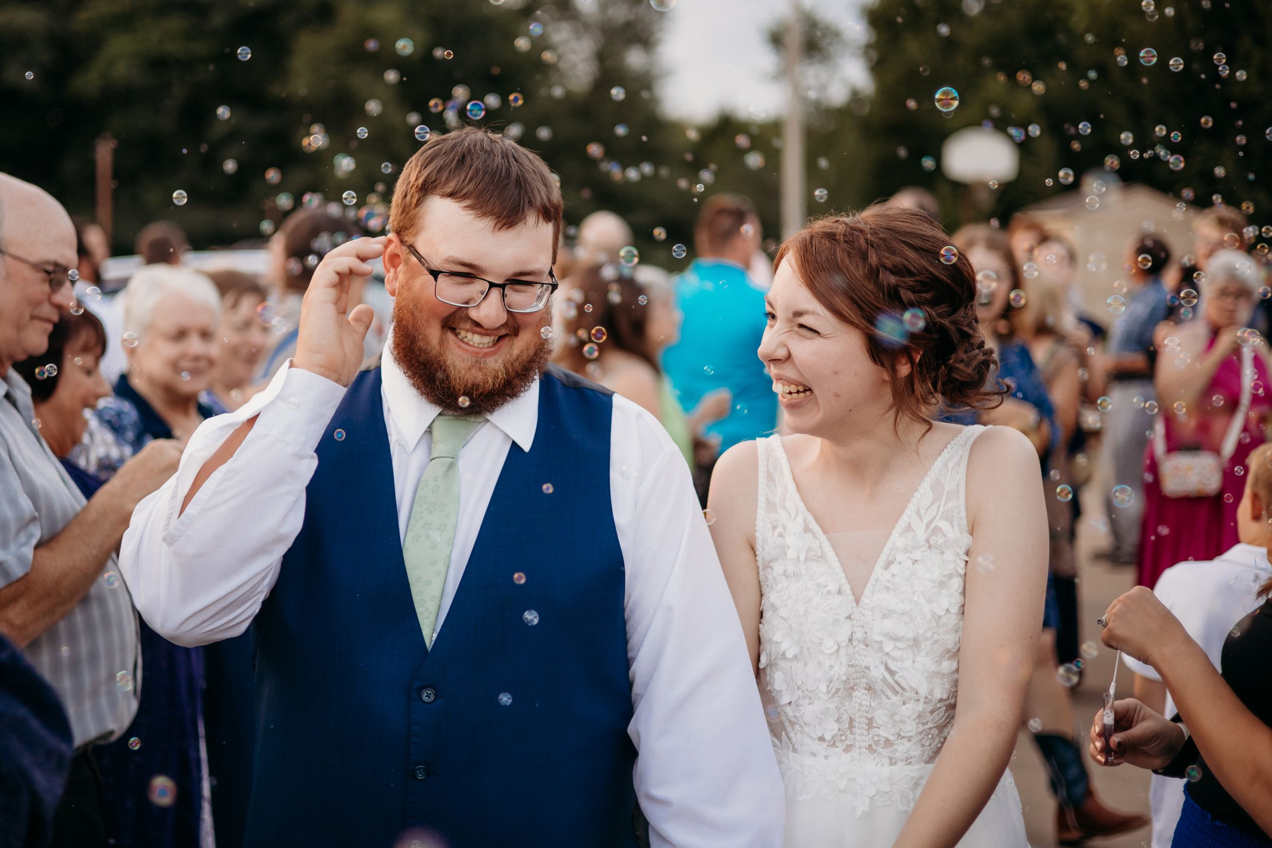 A groom and bride hold hands and giggle while departing from their wedding reception to a parade of bubbles and cheers from their loved ones