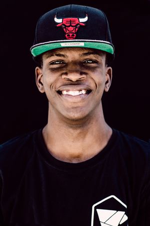 Smiling young man wearing a Chicago Bulls snapback cap and black t-shirt against a dark background.