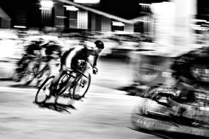 Black and white motion blur photo of cyclists racing at high speed through a city street at night.