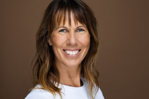Professional headshot of someone wearing a white top and smiling warmly against a brown background.