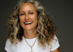 Portrait of someone with curly blonde hair wearing a white t-shirt and necklace, laughing joyfully against dark background.