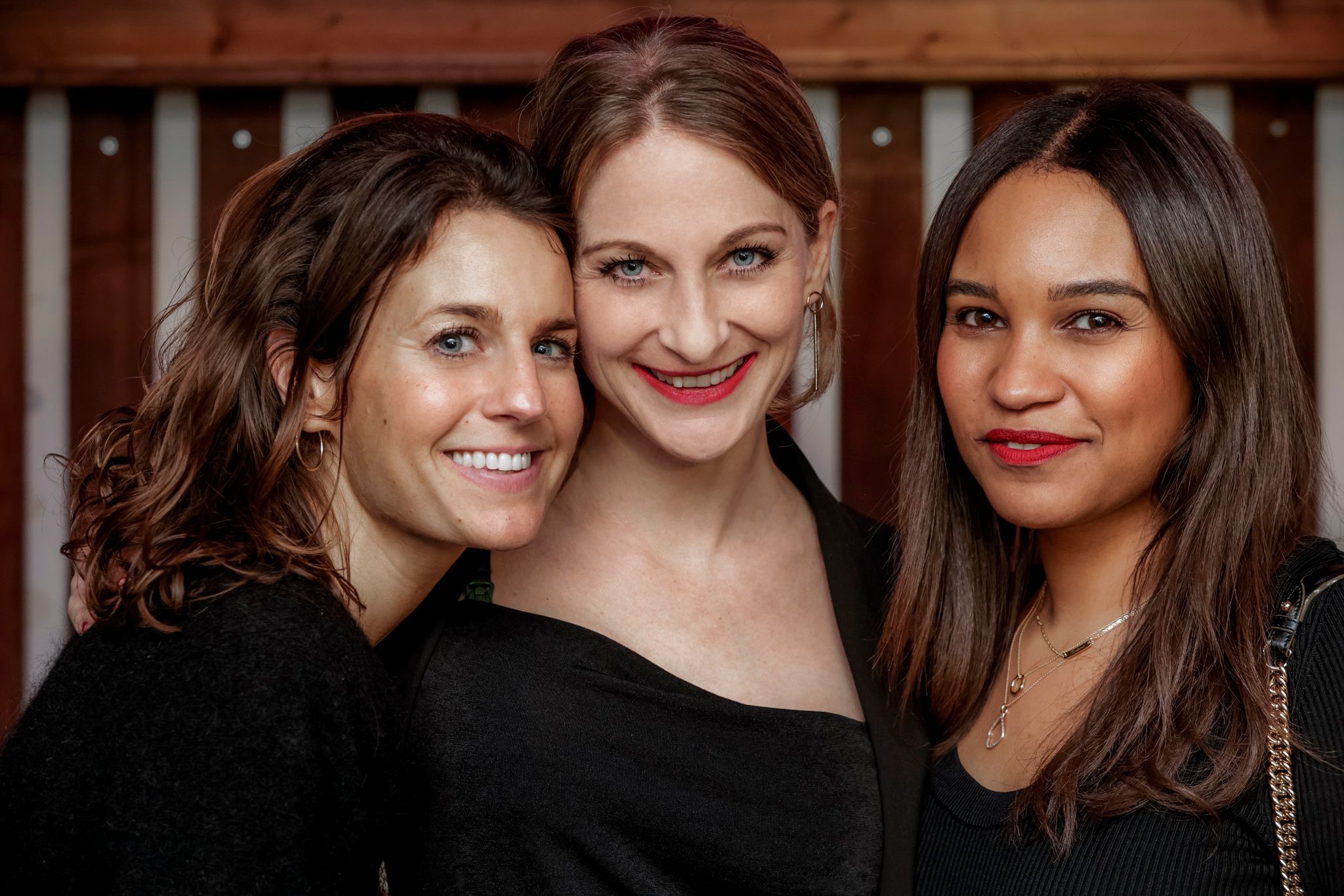 Three smiling women in black tops posing closely together indoors for a friendly group portrait photo.