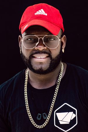 Studio portrait of person wearing red Adidas cap, glasses and gold chain against black background.