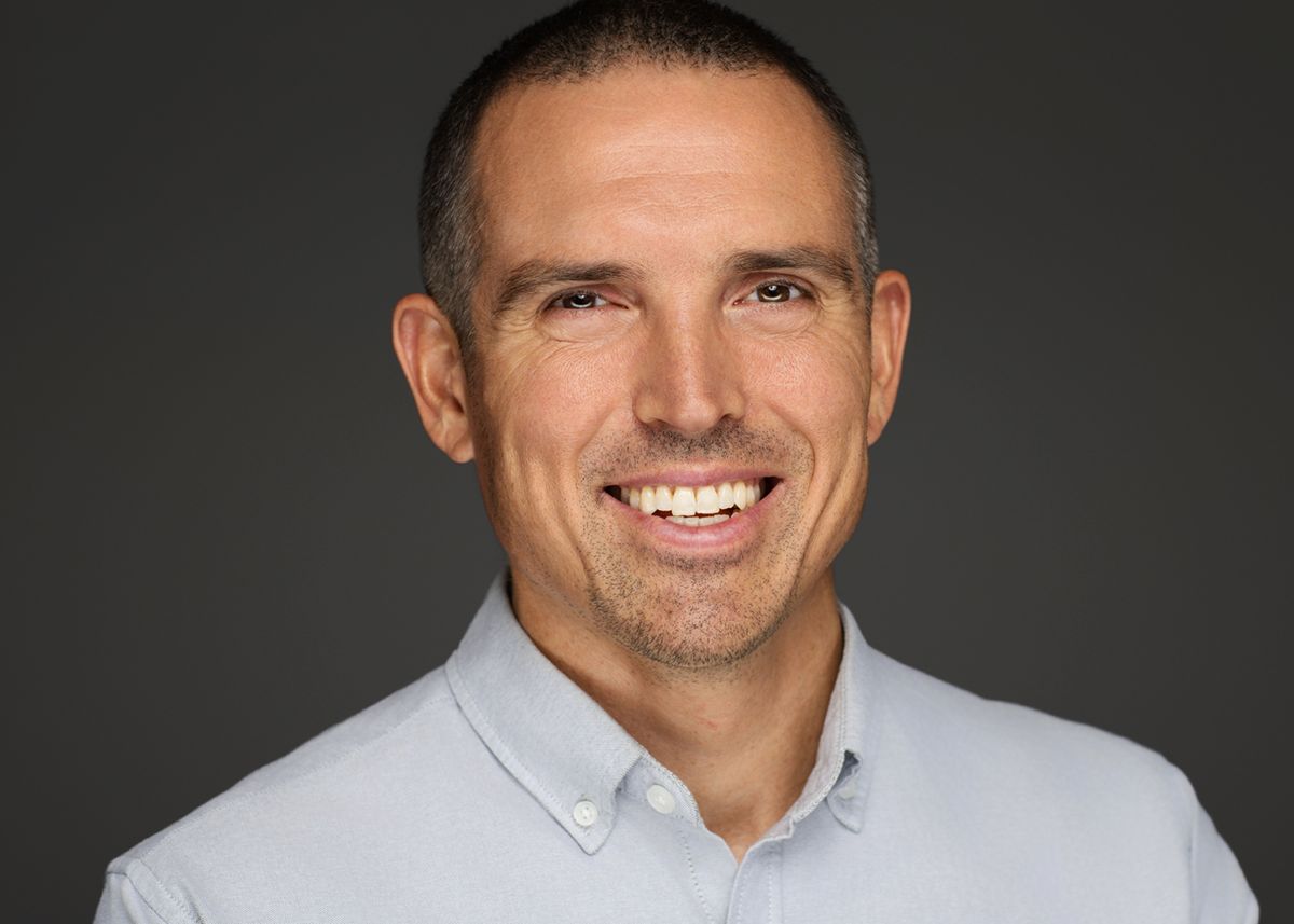 Professional headshot against dark background wearing light gray collared shirt with bright warm smile.