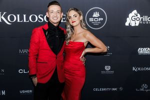 Couple in matching red formal attire posing at glamorous event with sponsorship backdrop.