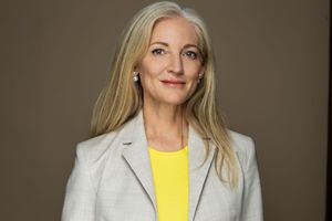 Professional woman with blonde hair wearing a yellow top and light gray blazer poses for a corporate headshot.