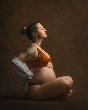 Artistic maternity portrait in warm tones showing expectant mother in orange top sitting peacefully in a meditative pose.
