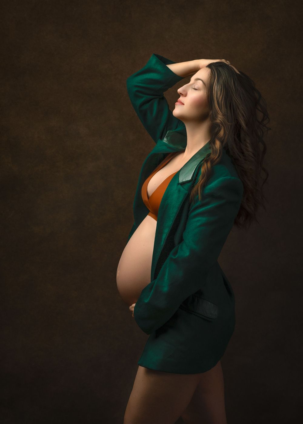A beautiful maternity portrait shows a model in emerald green blazer and orange bra against a dark backdrop.