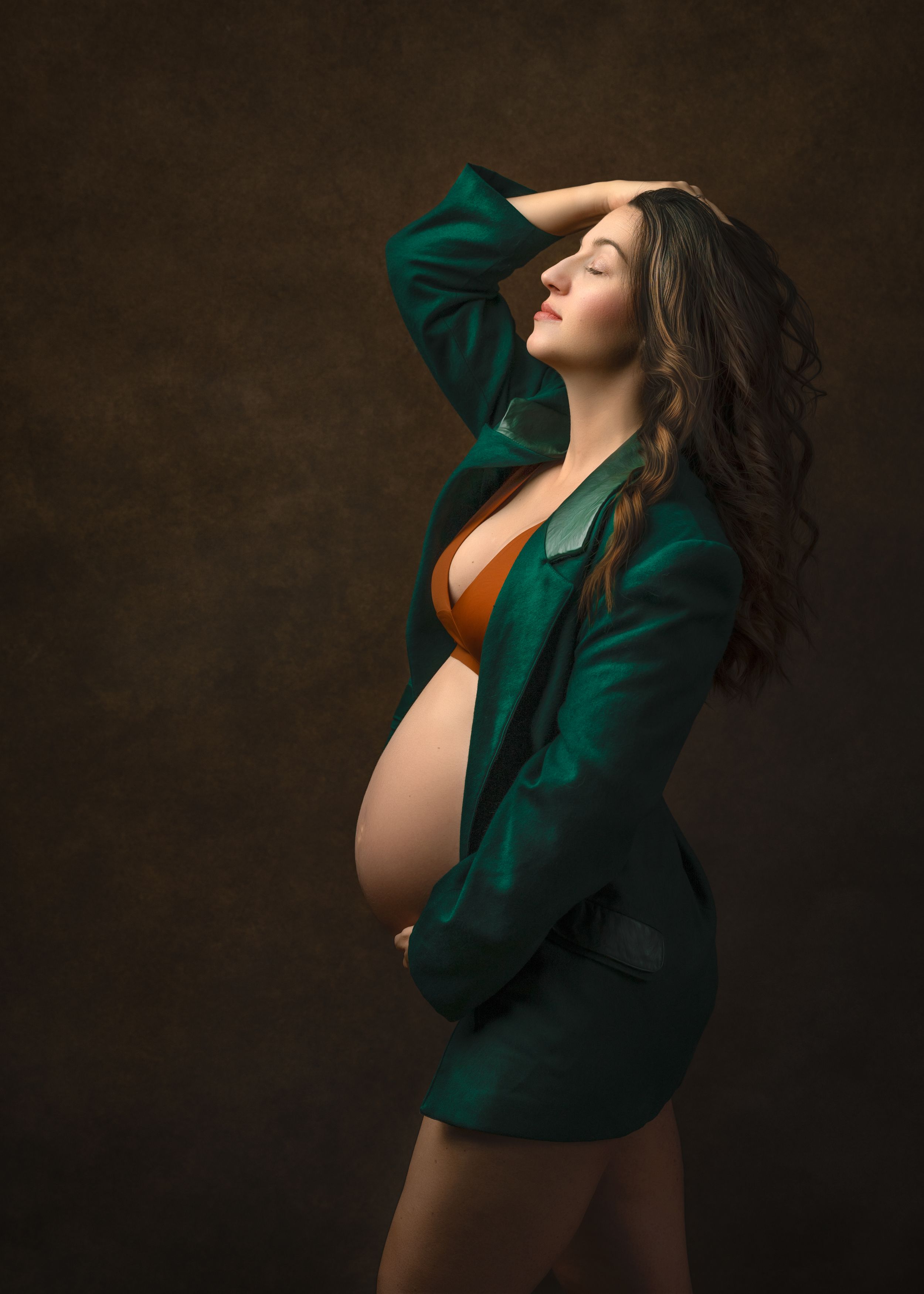 A beautiful maternity portrait shows a model in emerald green blazer and orange bra against a dark backdrop.