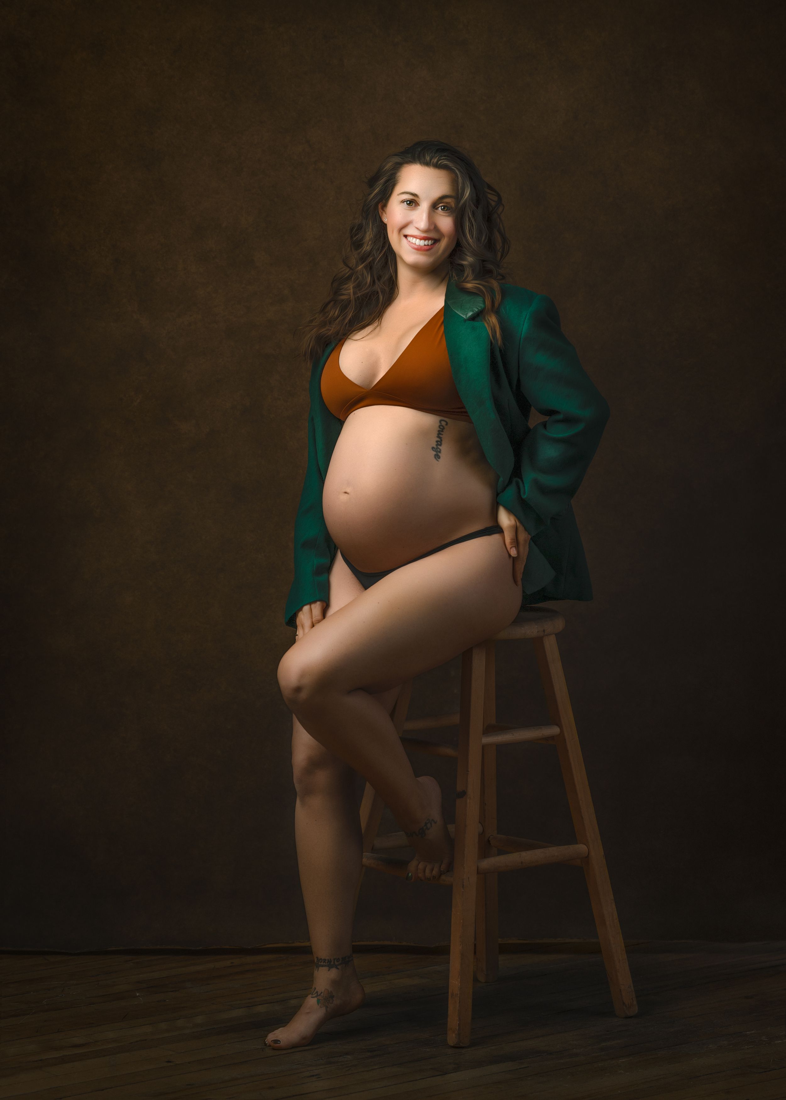 A pregnant figure in a rust-colored bra and emerald green jacket poses elegantly on a wooden stool against a dark background.