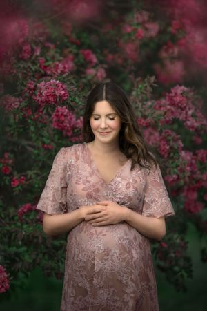 Expectant mother in pink lace dress standing before flowering pink bushes.