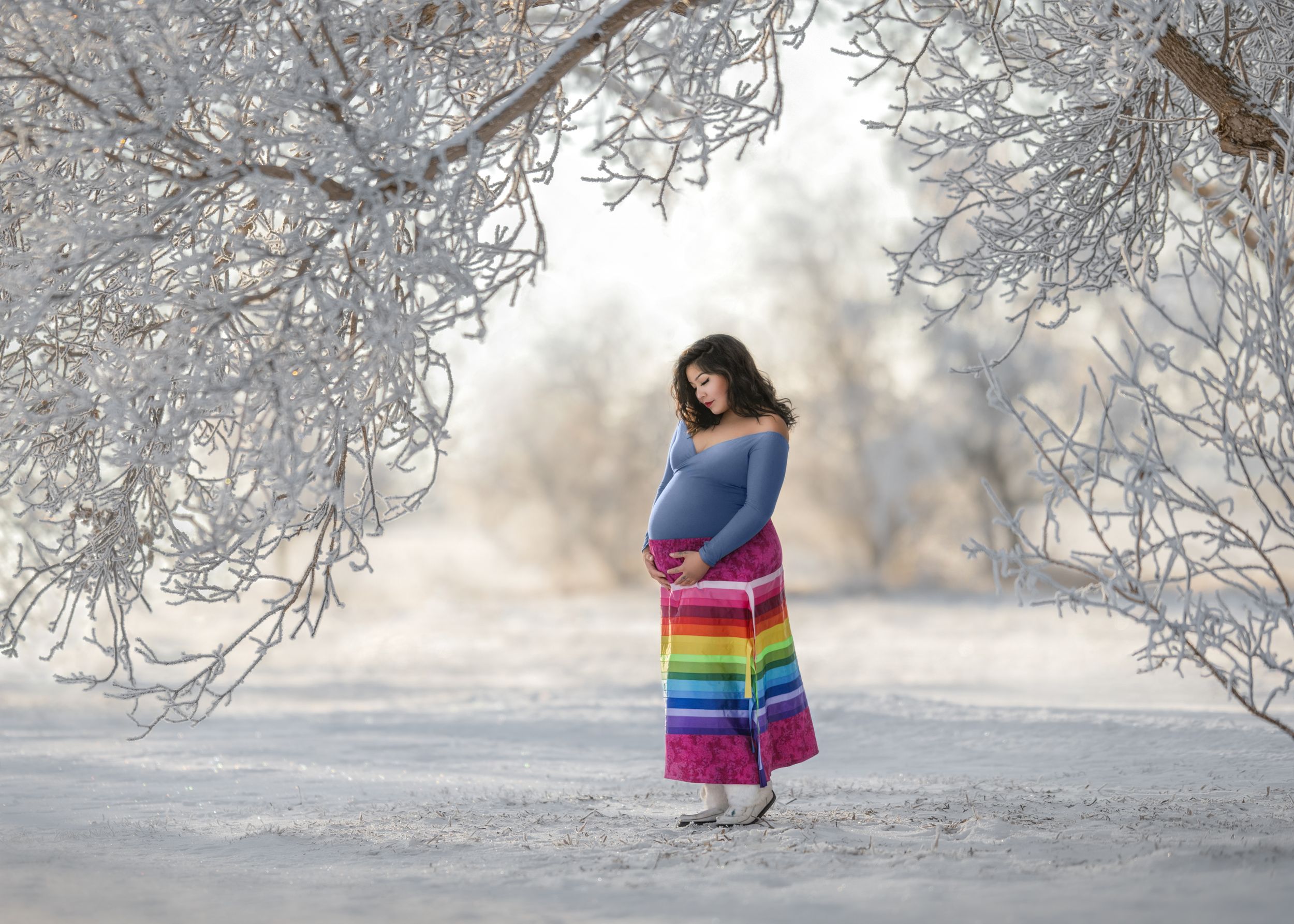 Maternity portrait featuring a lady in an Indigenous rainbow ribbon skirt on a winter landscape with frosted trees in Saskatchewan, Canada