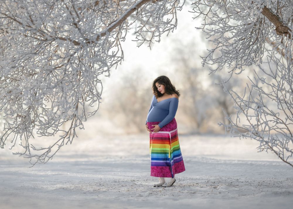 Maternity portrait featuring a lady in an Indigenous rainbow ribbon skirt on a winter landscape with frosted trees in Saskatchewan, Canada
