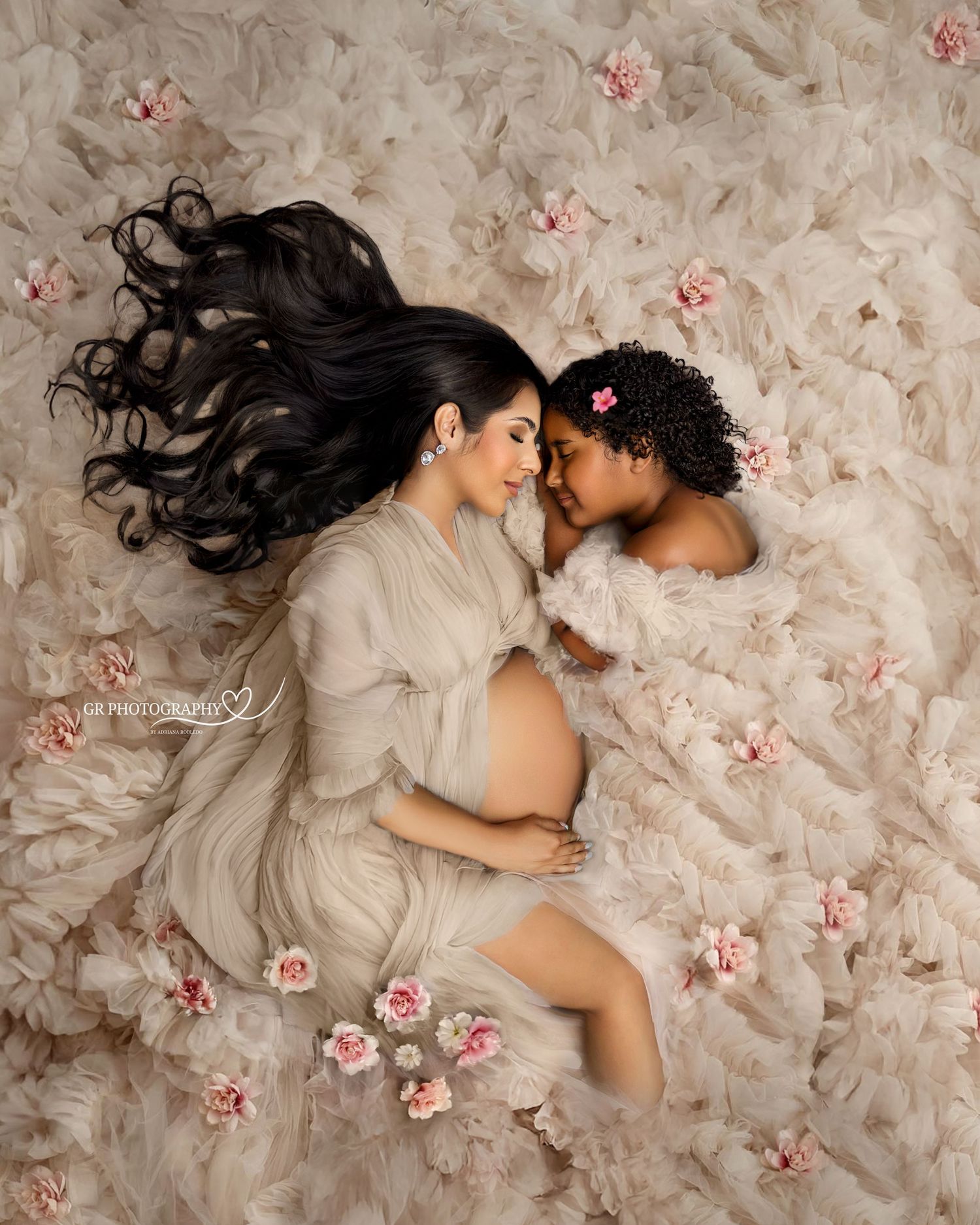 Pregnant woman in sheer gown lying with young girl among pink roses on soft cream ruffled fabric in maternity photo.