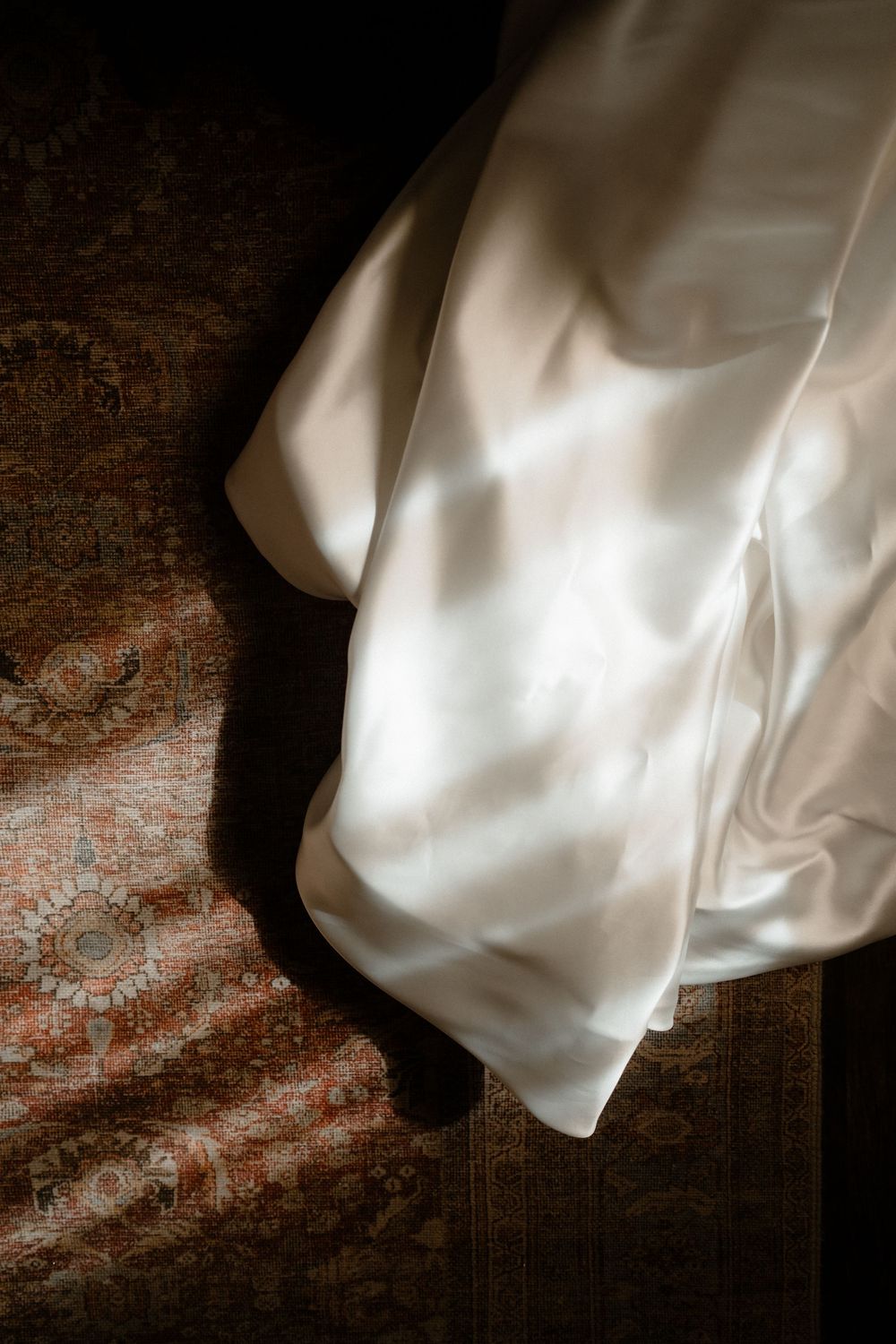 White satin wedding dress draped over an ornate patterned rug in dramatic natural lighting.