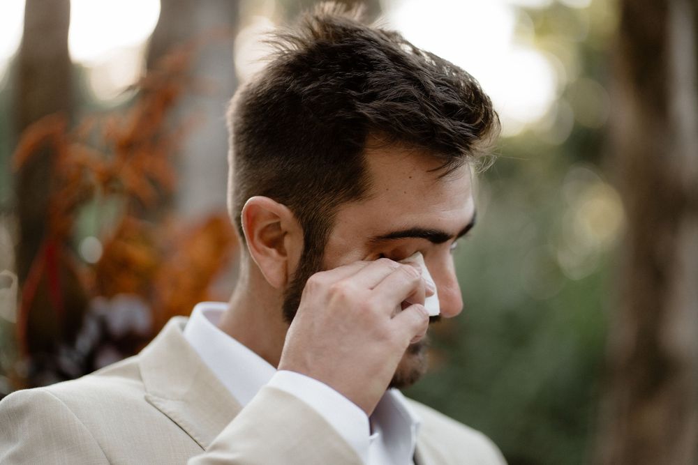 Emotional groom wiping a tear during his outdoor wedding ceremony wearing a beige suit.