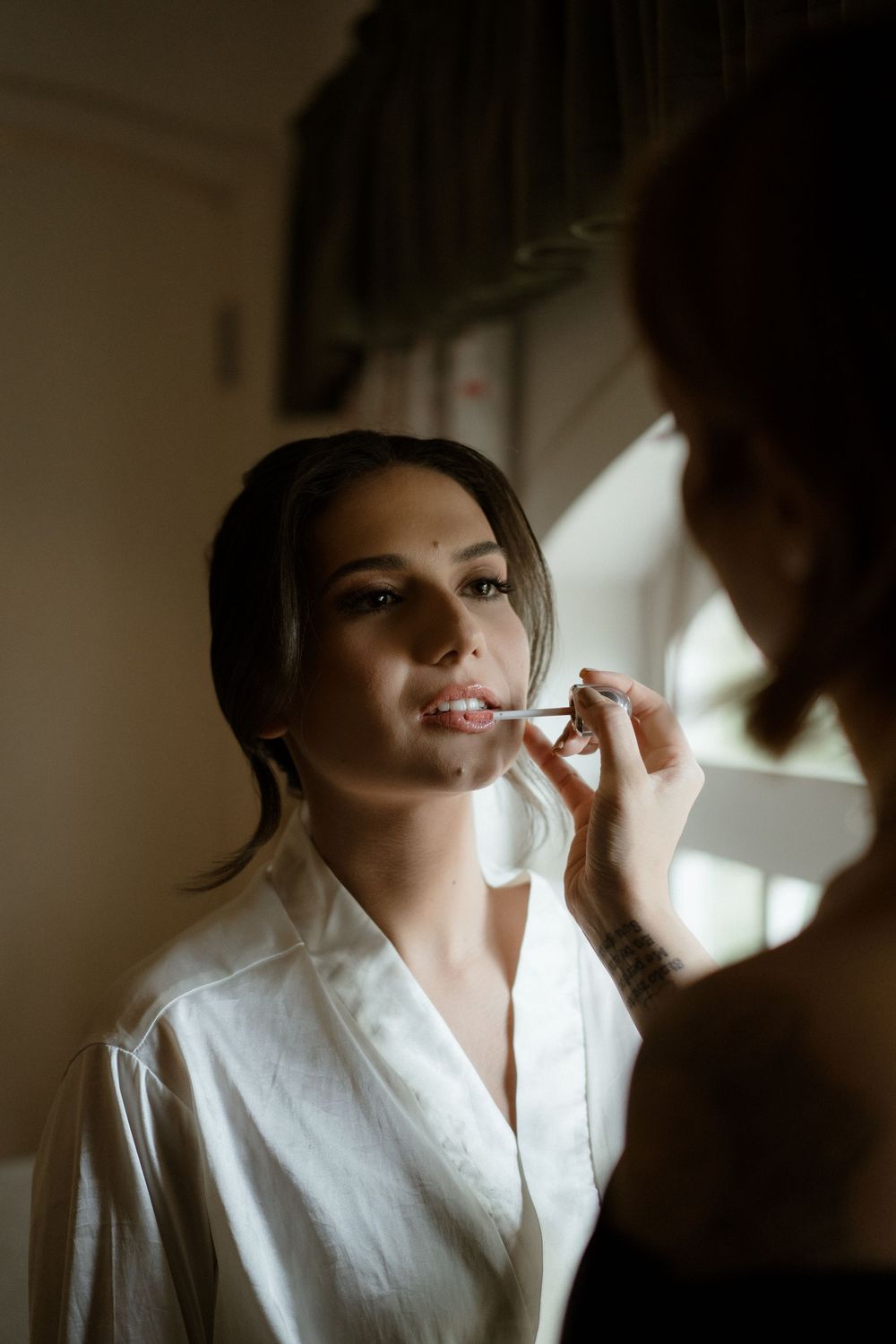 A woman in a white collared shirt gets her makeup applied in a series of soft, naturally lit photos.