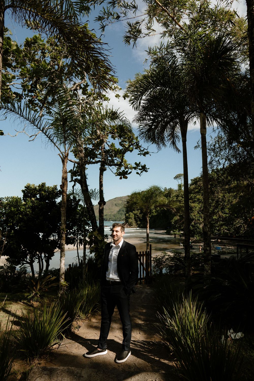 Groom in black suit standing among tropical palm trees with ocean and hills in background on wedding day.