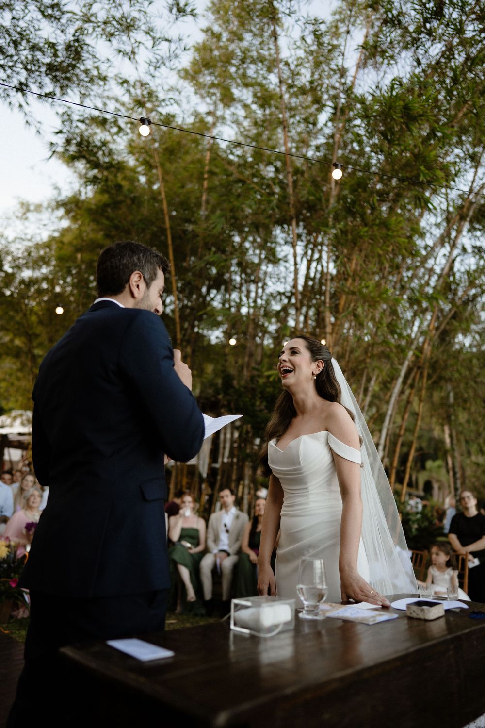 Bride laughing joyfully at outdoor wedding ceremony while groom reads vows, surrounded by trees and string lights.