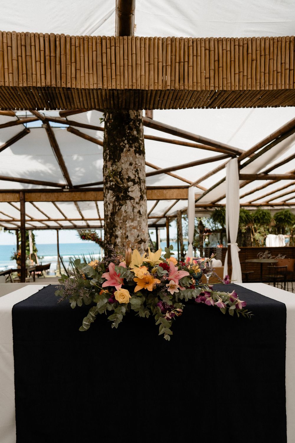 Tropical beach venue with bamboo roof, tree trunk centerpiece, and colorful floral arrangement on black draped table.