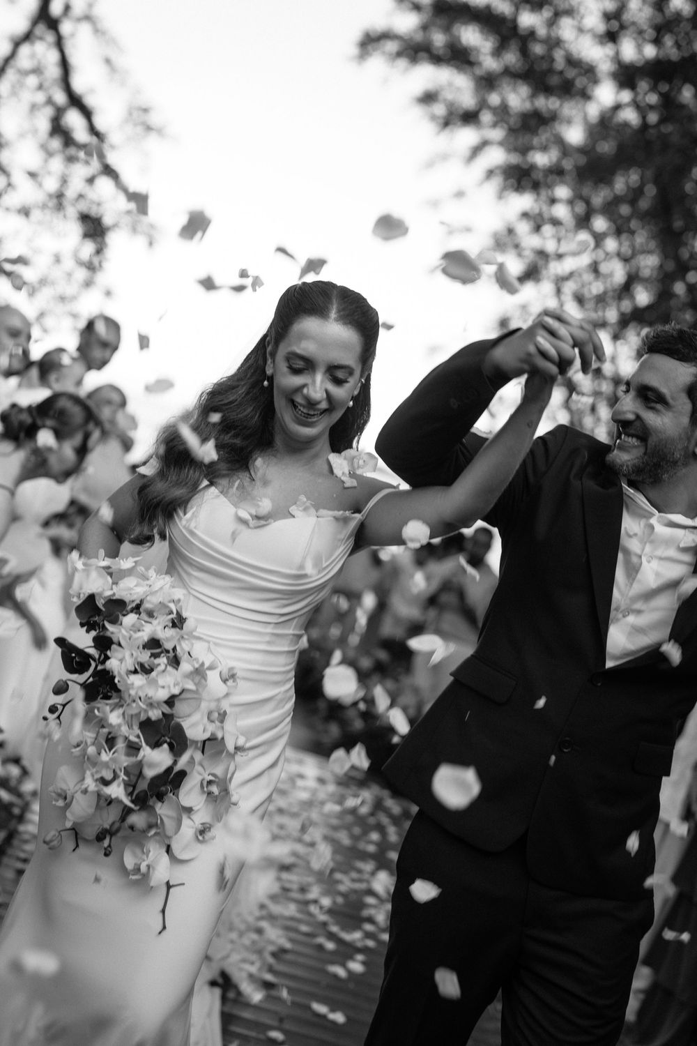 Black and white photo of a joyful bride and groom walking through a shower of flower petals at their wedding.