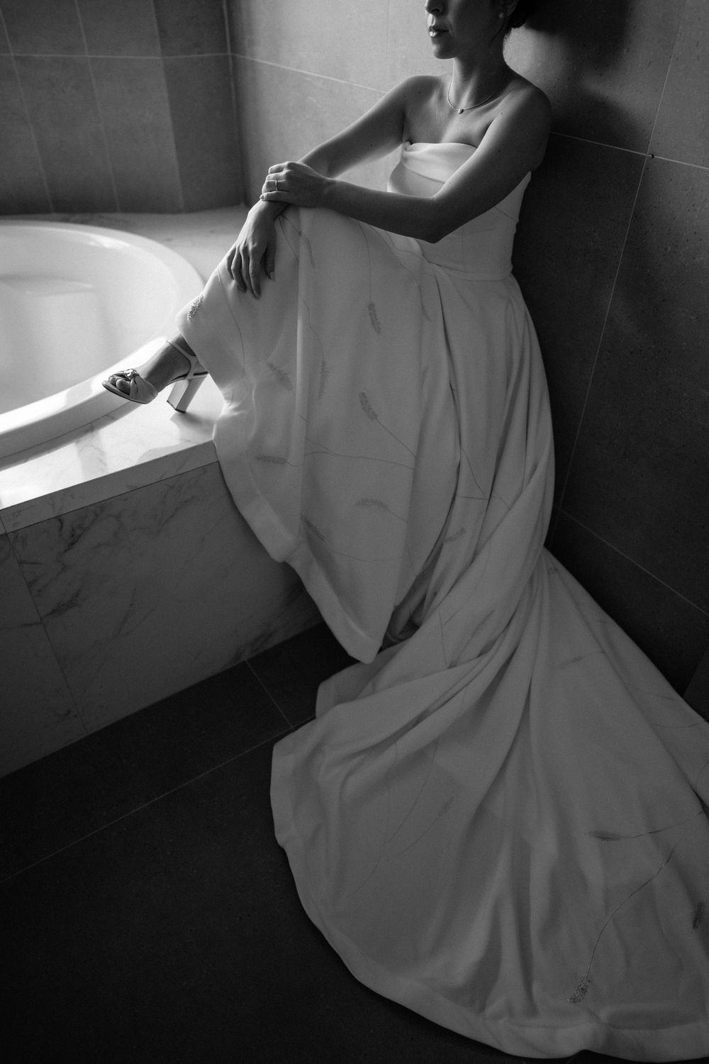 Black and white photo of a bride in a white gown seated elegantly beside a marble bathtub.