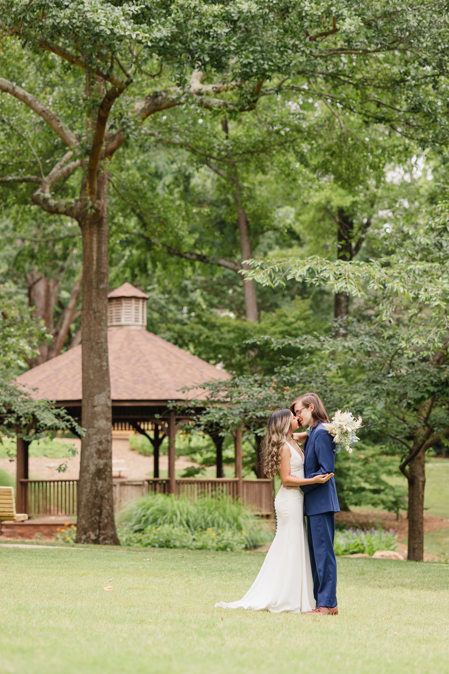 A couple embraces in their wedding attire in front of a wooden gazebo surrounded by lush green trees.