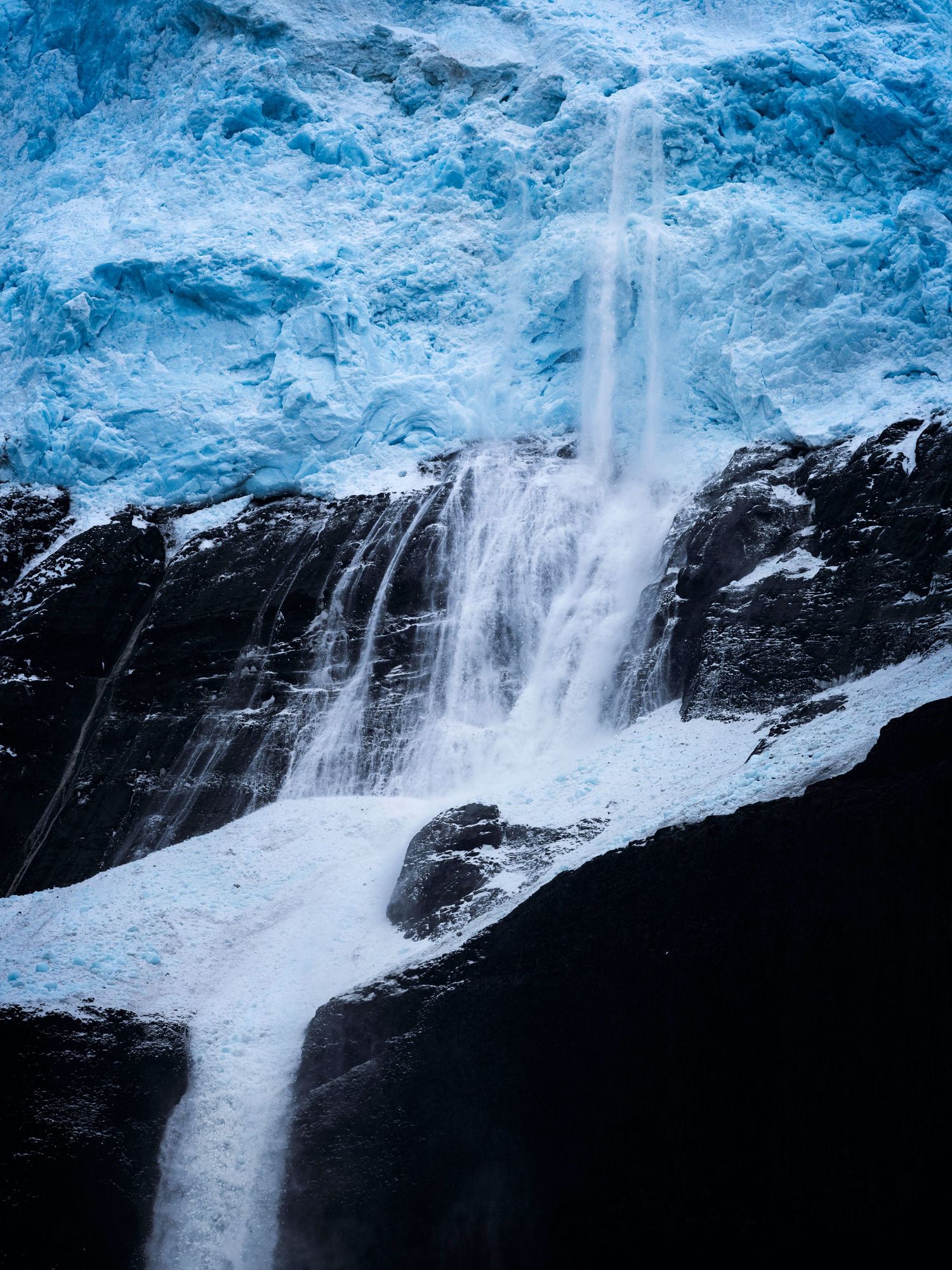 Dramatic waterfall cascading down dark rocky cliffs with blue glacial ice above.