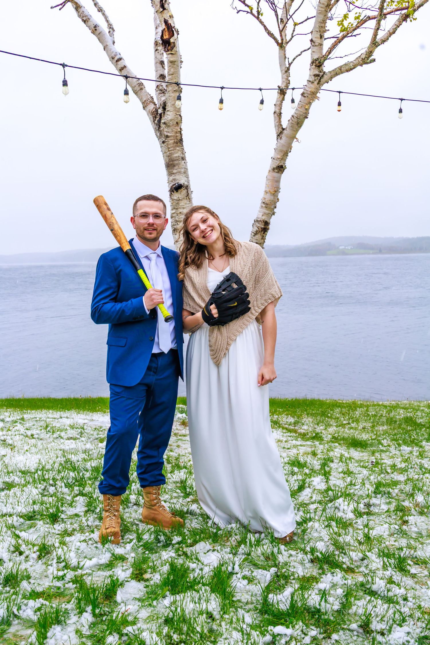 Bride and groom posing outdoors by waterfront, holding baseball bat and glove in wedding attire near birch trees.