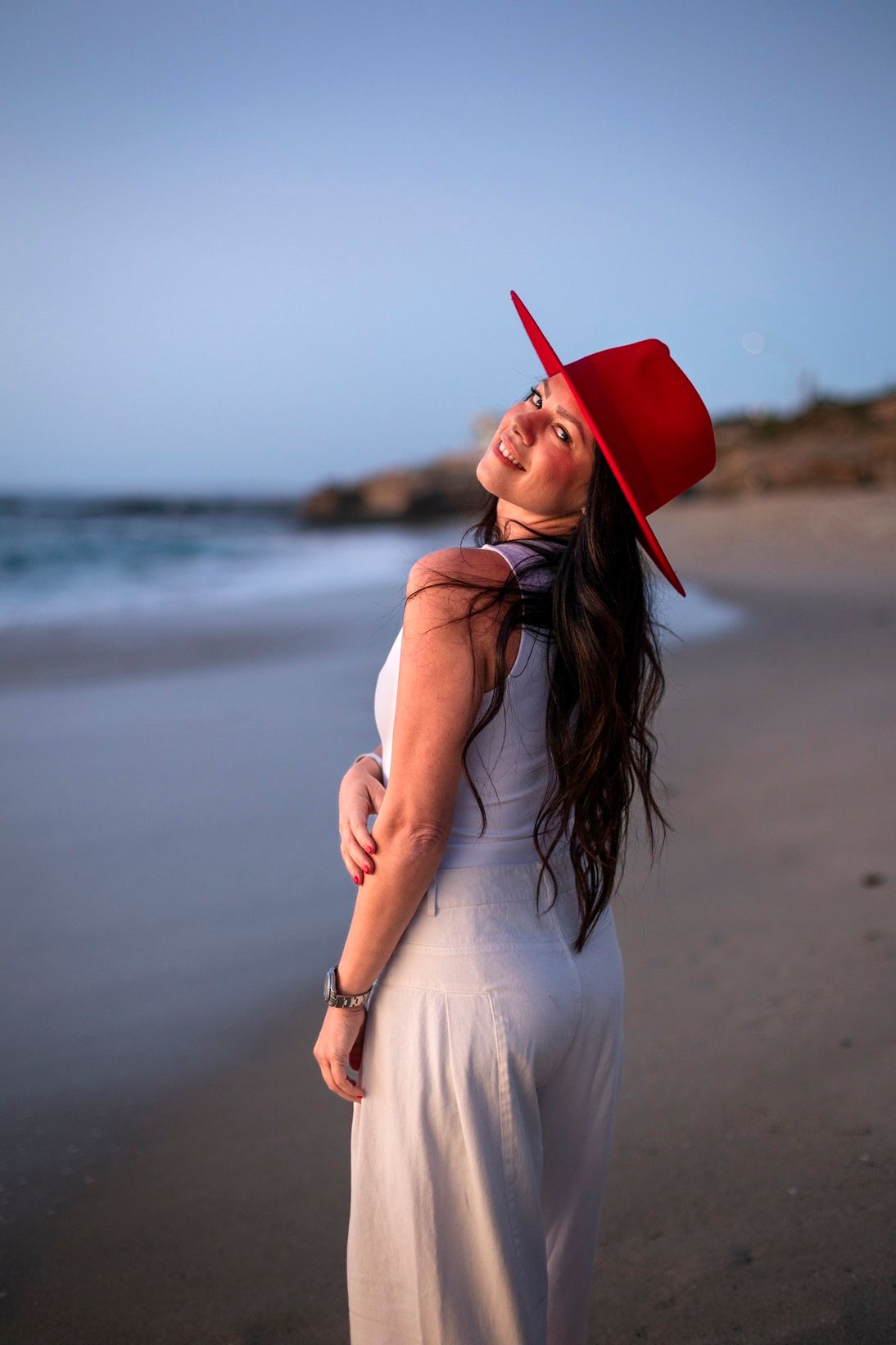 Woman in white dress and red hat poses gracefully on a sandy beach at sunset.