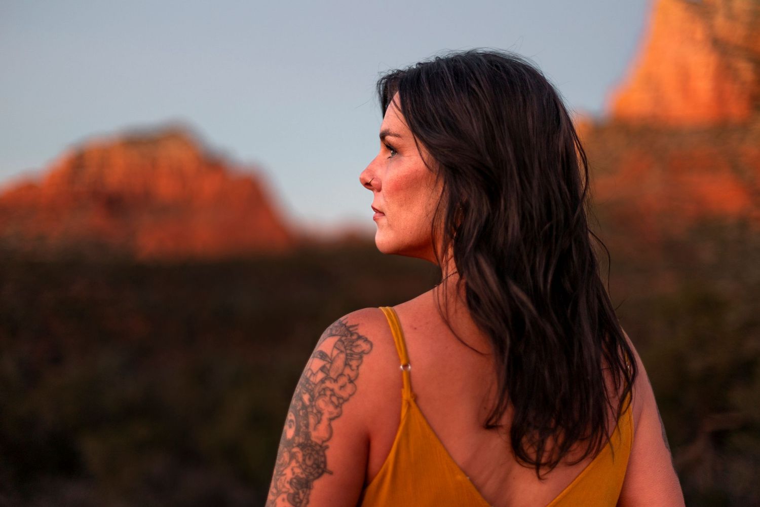 Woman with tattoo in yellow top gazes at red rock formations during golden hour in Sedona, Arizona.